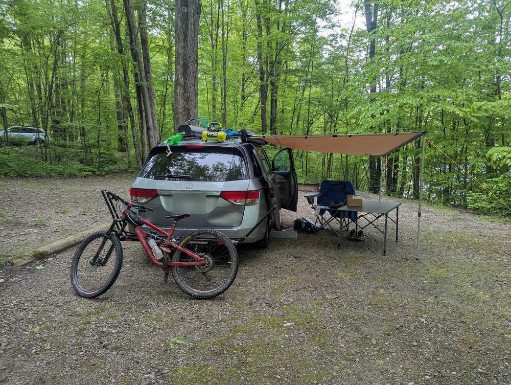 A campsite in the woods with an awning off the van and a muddy mountain bike on the rack